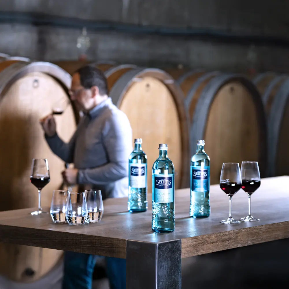 Vier Frauen sitzen lachend an einem Tisch in einem Weinberg und trinken Getränke.
