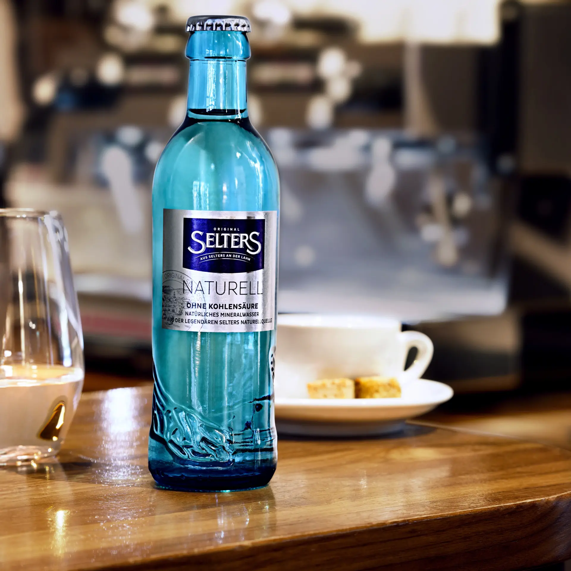 Eine blaue Mineralwasserflasche steht auf einem Holztisch, im Hintergrund eine Tasse und ein Glas.