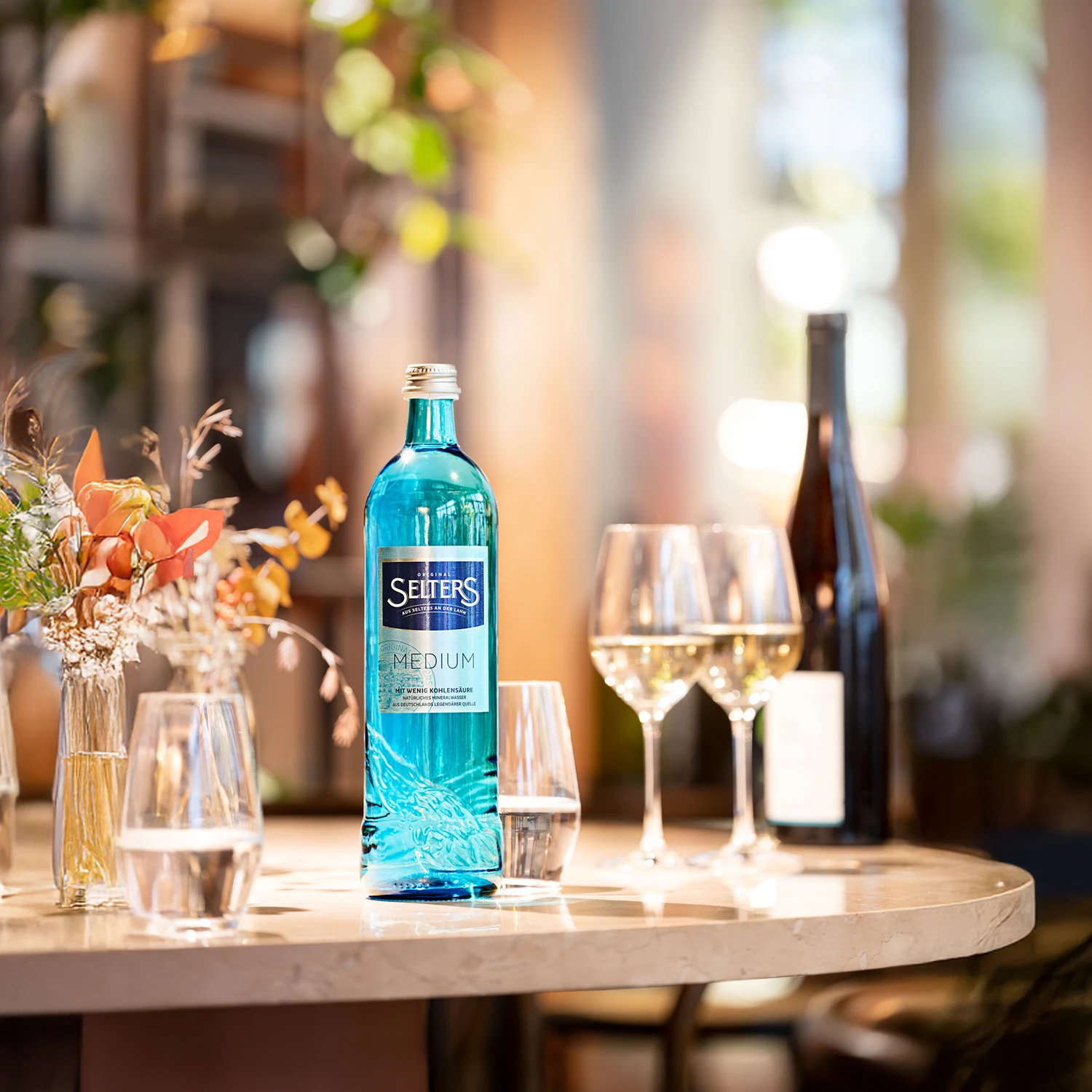 Eine blaue Wasserflasche steht auf einem Tisch umgeben von Weingläsern und Blumen. Hintergrund unscharf mit Lichtspielen.