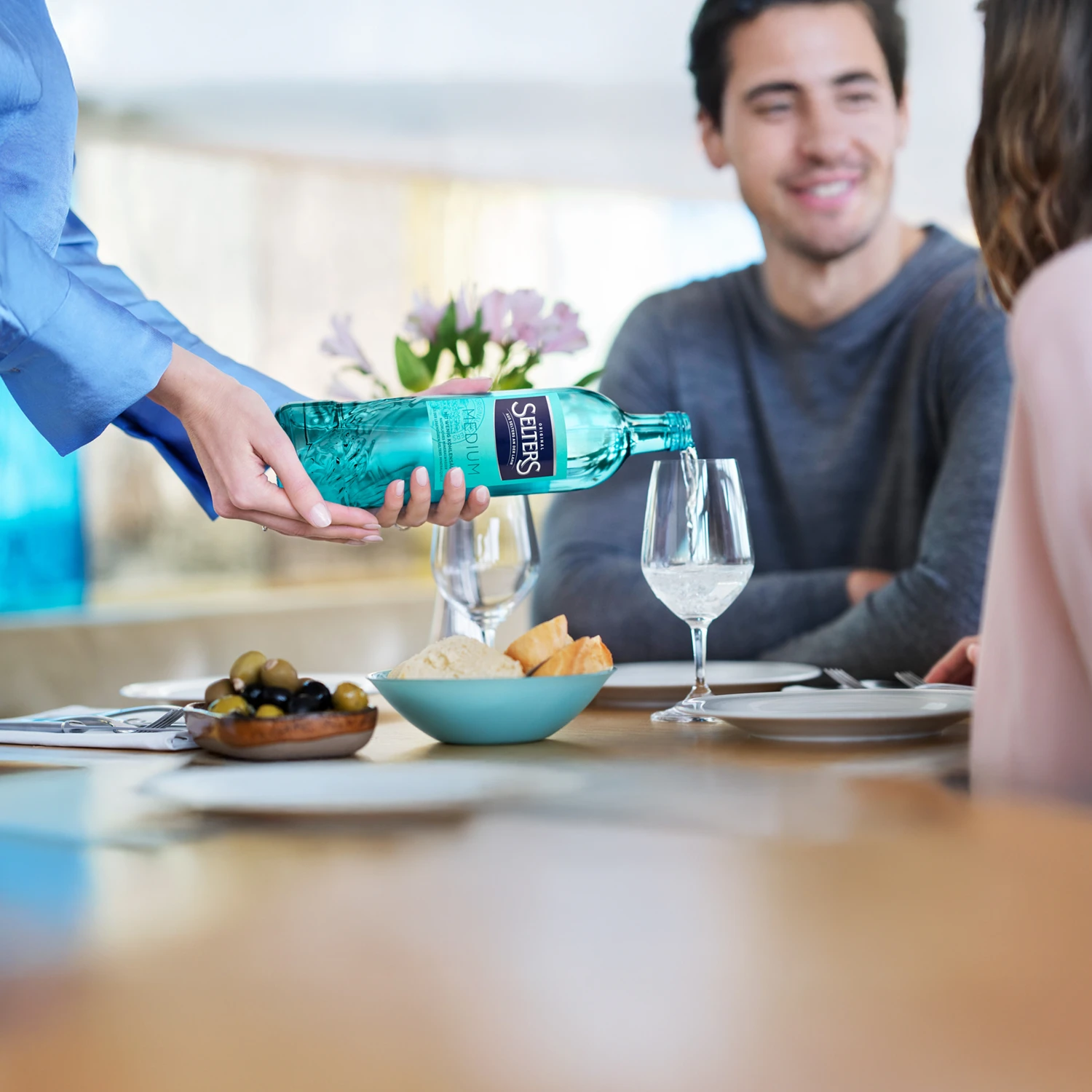 Eine Bedienung gießt Wasser aus einer grünen Flasche in ein Weinglas, während ein Paar am Tisch sitzt.