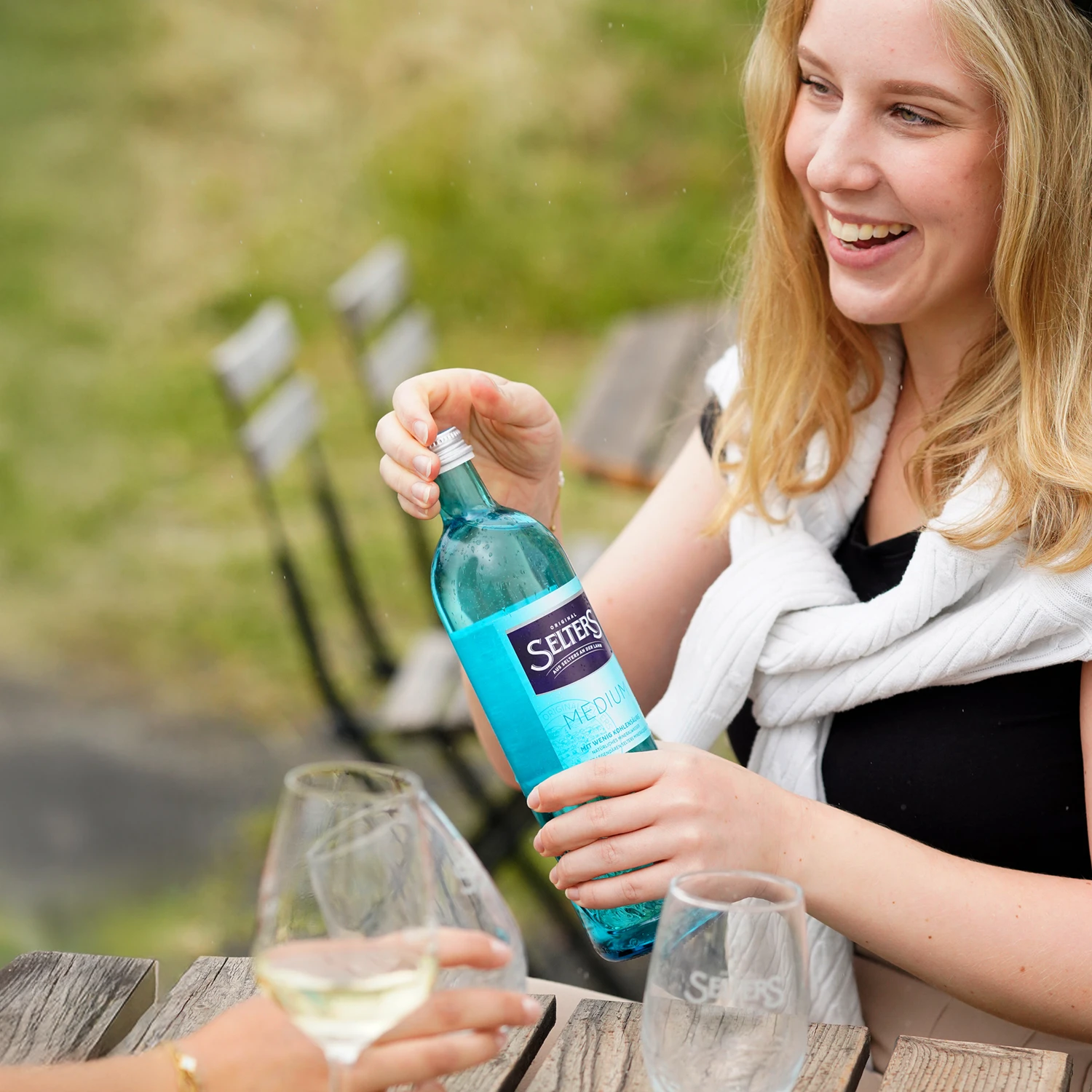Eine lächelnde junge Frau füllt ein Glas mit Mineralwasser an einem Holztisch im Freien.