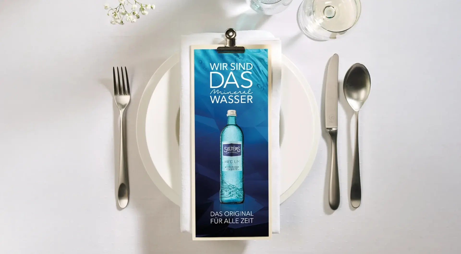 Ein gedeckter Tisch mit Besteck und einer Flaschenwerbung in der Mitte. Die Werbung liegt auf einer Serviette.