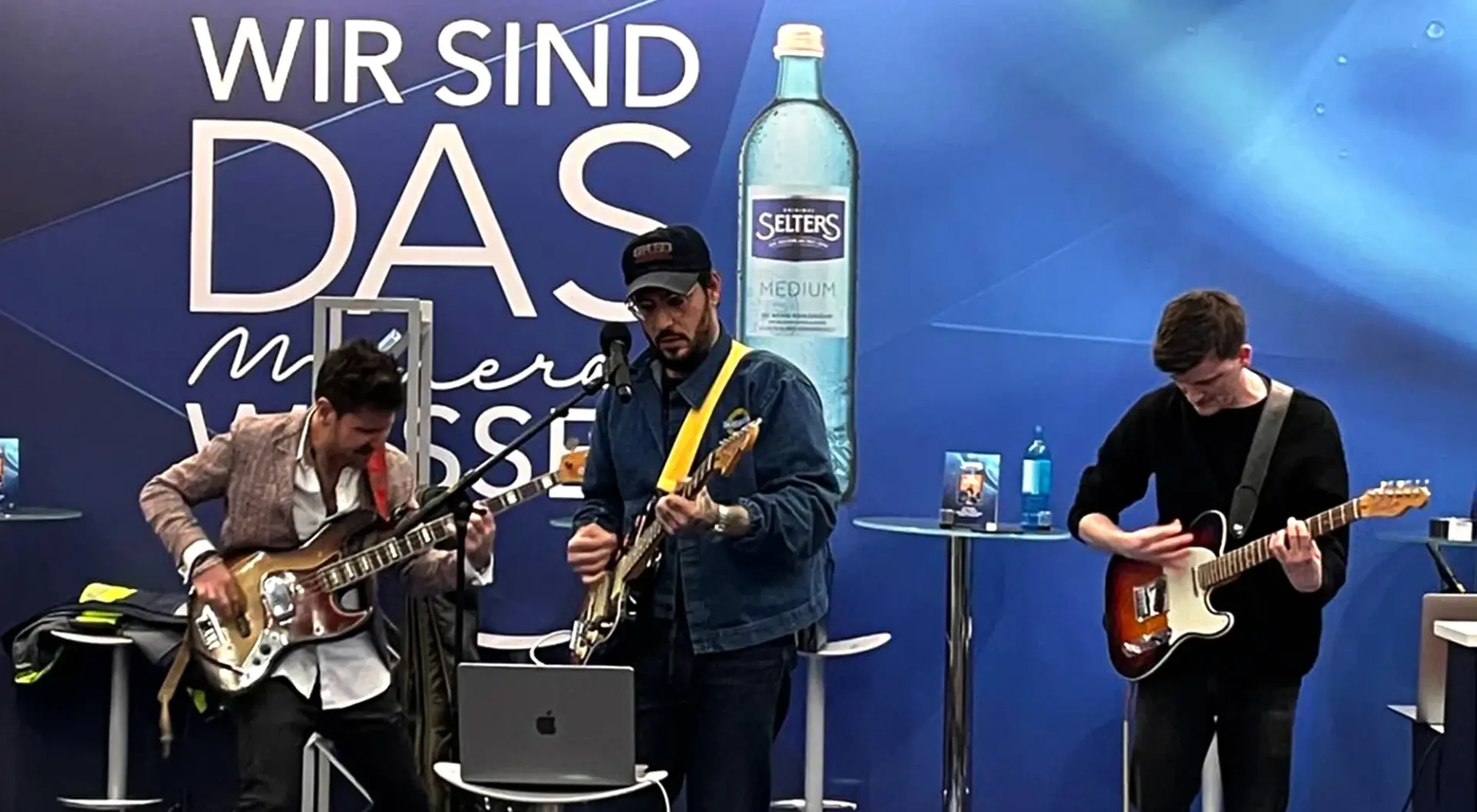 Drei Musiker spielen Gitarre auf einer Bühne vor einem blauen Hintergrund.